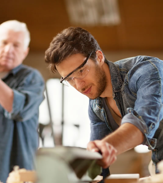 Young handyman wearing safety glasses working with tools while an older man observes in the background