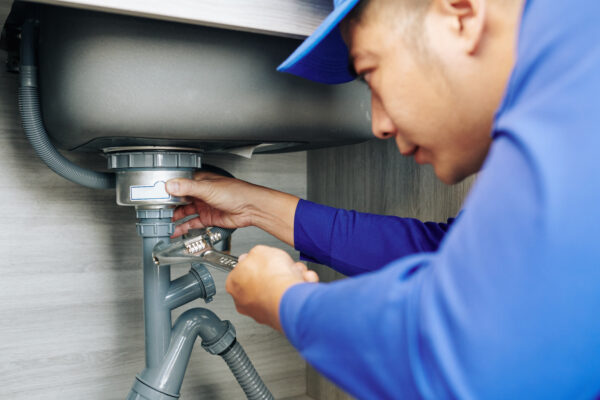 Professional plumber adjusting a kitchen sink drain and P-trap with an adjustable wrench beneath the counter.