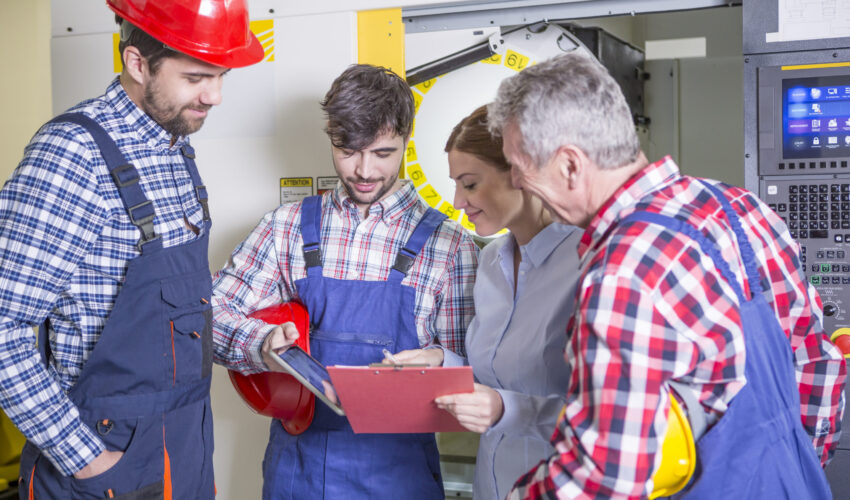 Technicians in uniforms reviewing a checklist beside industrial machinery at a Maintenance Company in Dubai, discussing equipment safety and repair tasks.