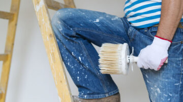 Painter holding a large brush while standing on a ladder with paint splattered jeans.