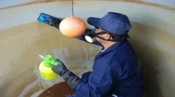 Technician in safety gear spraying cleaning solution inside a water tank.